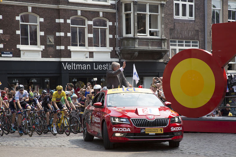 Tourpeloton dat de Stadhuisbrug te Utrecht oprijdt voorafgegaan door de auto van de Tourleiding met daarin o.a. burgemeester mr. J.H.C. van Zanen tijdens de start van de tweede etappe van de Tour de France 2015